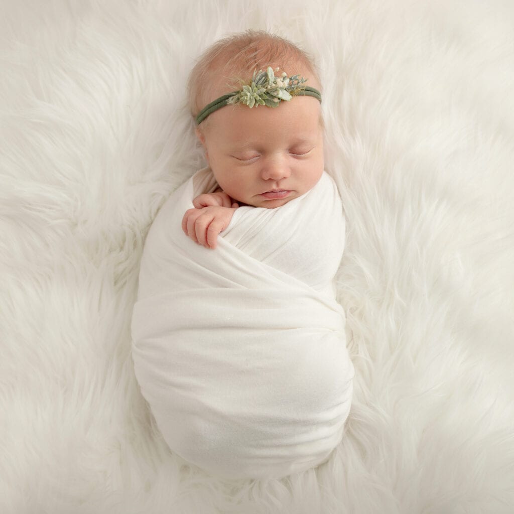 Nyfødt baby sovende i hvit pose med blomsterhodebånd.