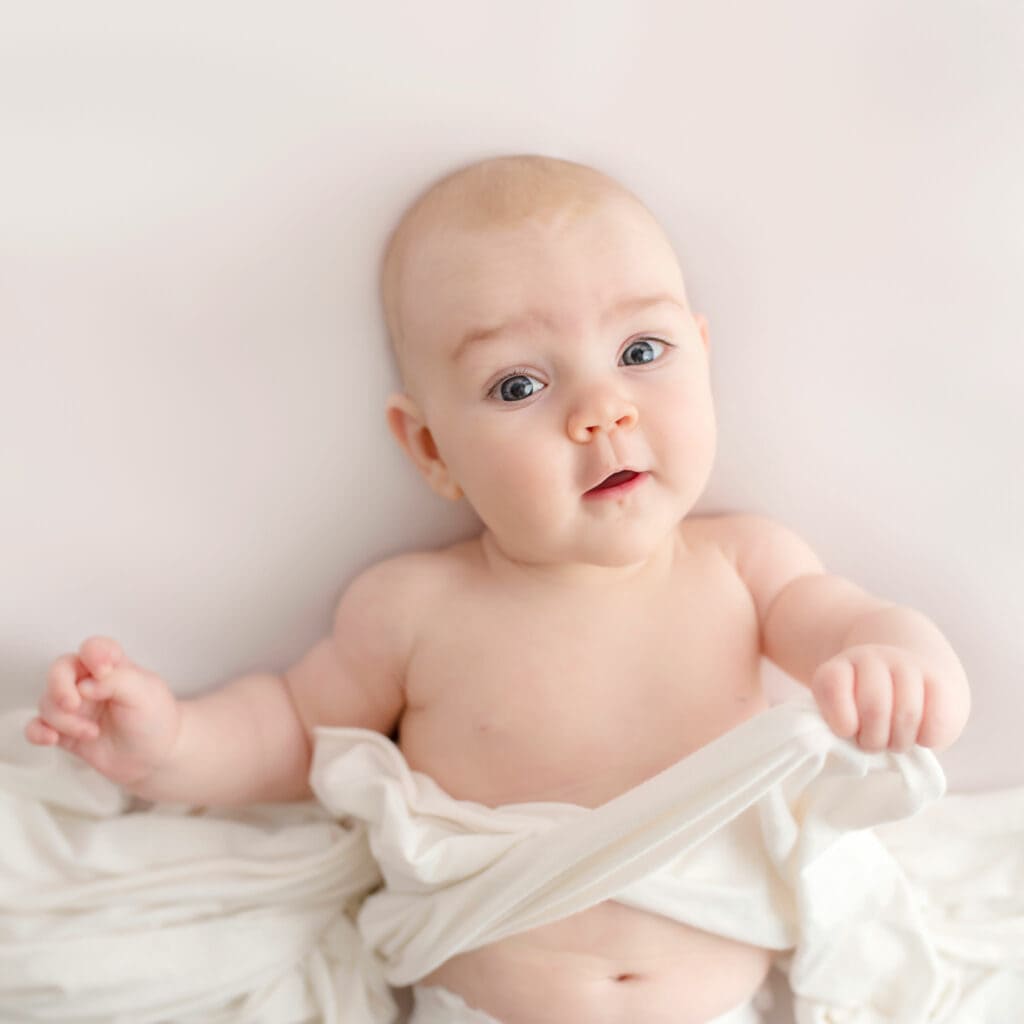 Baby med blå øyne og lys hud som ligger på seng, ser nysgjerrig ut.