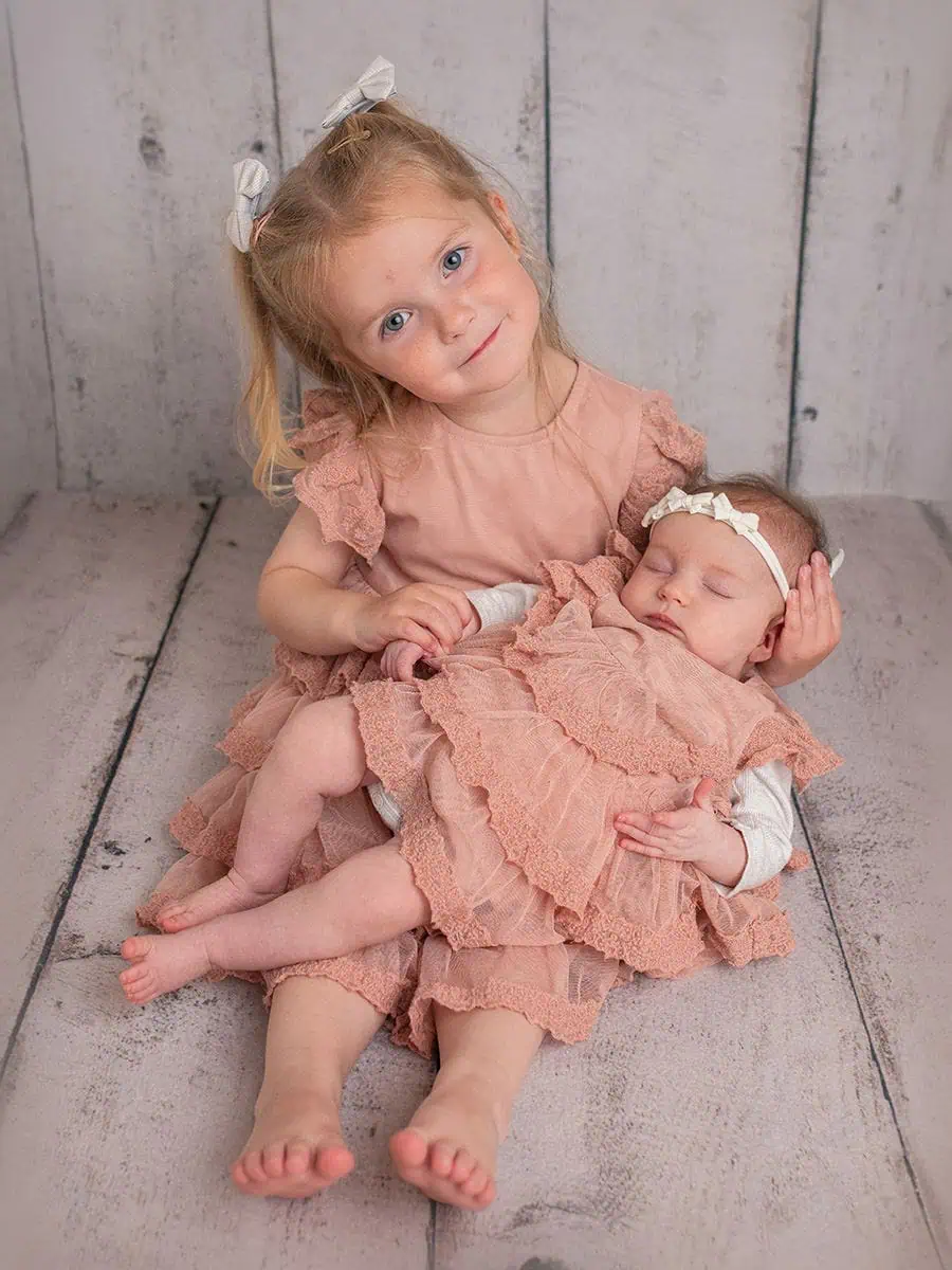 Søster holder nyfødt baby, begge i rosa klær, i en fotoshoot.