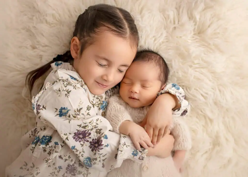 Storesøster holder nyfødt baby tett inntil seg på mykt teppe i studio i Jessheim