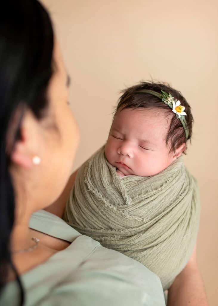 Nyfødt baby i armene på mor, med blomsterhodebånd, i en varm og myk innendørs setting.